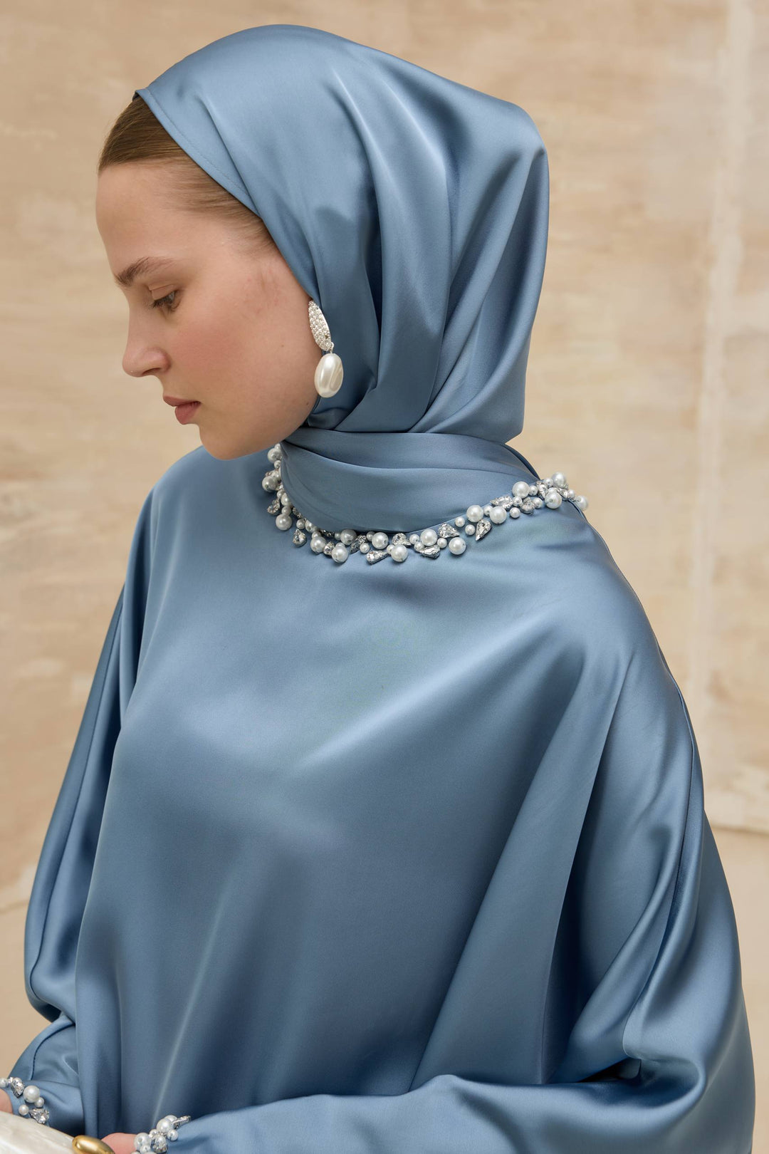 Woman wearing a blue hijab with pearl embellishments against a beige background