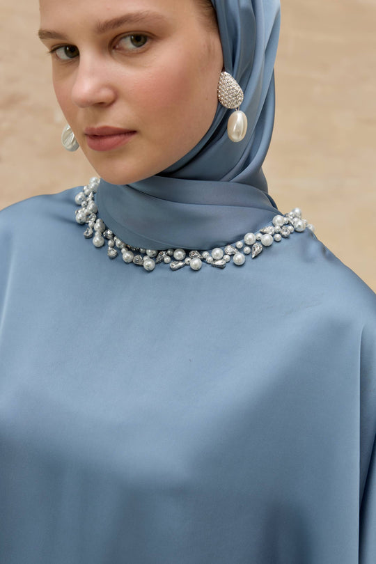 Woman wearing a blue hijab with pearl jewelry against a beige background