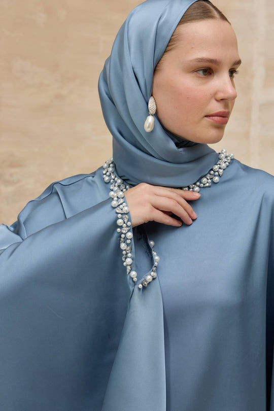 Woman wearing a light blue hijab and dress with pearl jewelry against a beige background