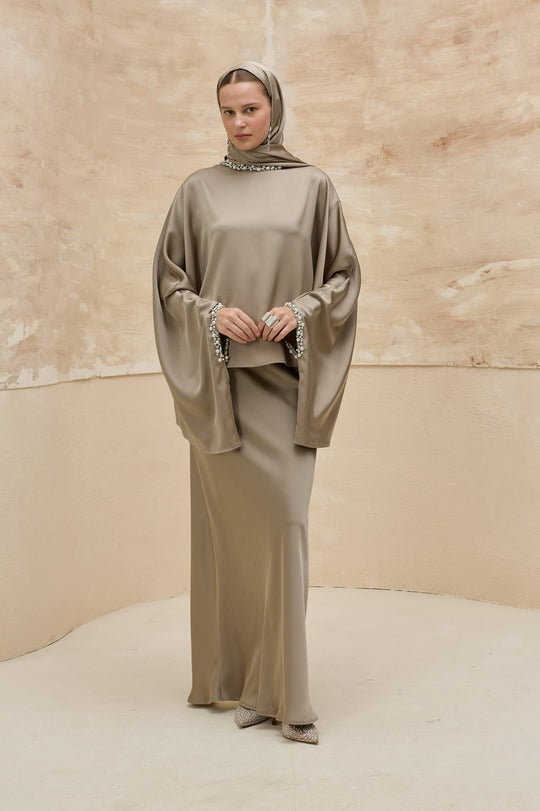 Woman wearing a long, light brown dress with a matching headscarf against a beige background