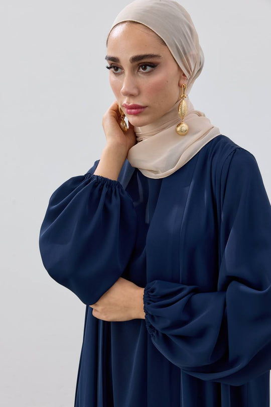 Woman wearing a navy blue dress with beige hijab against a light gray background