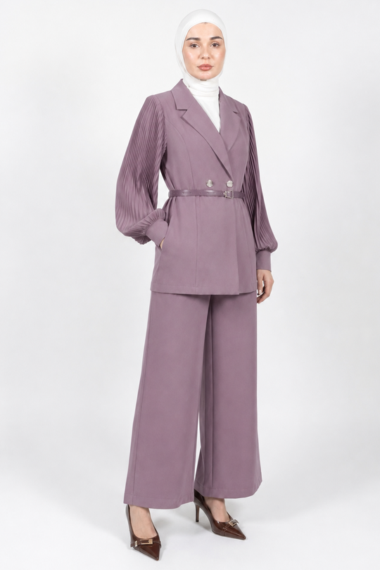 A woman wearing a matching mauve-purple two-piece suit with a double-breasted blazer and wide-leg trousers, styled with a white turtleneck, white hijab, and brown pointed-toe heels against a light gray studio background.