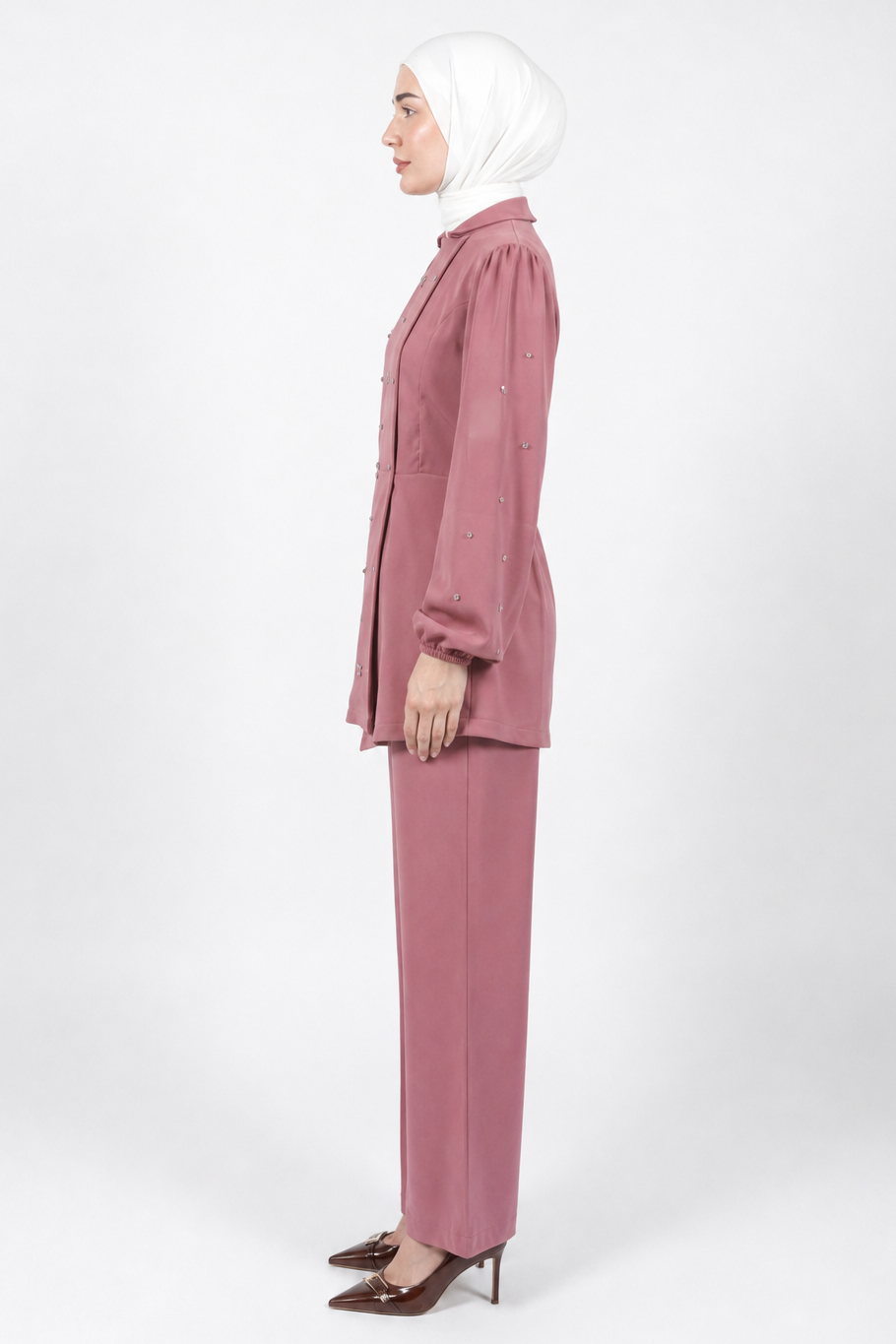 A woman in profile wearing a dusty rose two-piece suit with a long-sleeve button-up jacket and matching trousers, paired with a white hijab and brown pointed-toe heels, photographed against a seamless light gray studio background.