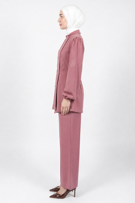 A woman in profile wearing a dusty rose two-piece suit with a long-sleeve button-up jacket and matching trousers, paired with a white hijab and brown pointed-toe heels, photographed against a seamless light gray studio background.
