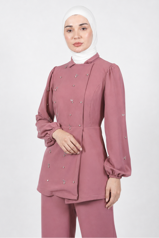 A woman wearing a dusty rose pink long-sleeve modest tunic top with matching pants and a white hijab, standing against a light gray studio background.
