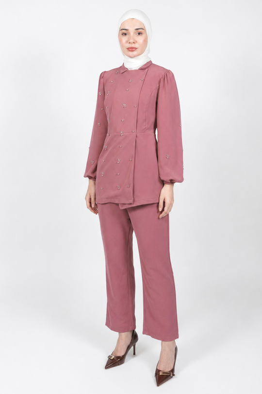 A woman modeling a dusty rose two-piece suit consisting of a long-sleeve button-front jacket and matching wide-leg pants, paired with a white hijab and brown pointed-toe heels, against a light gray studio background.