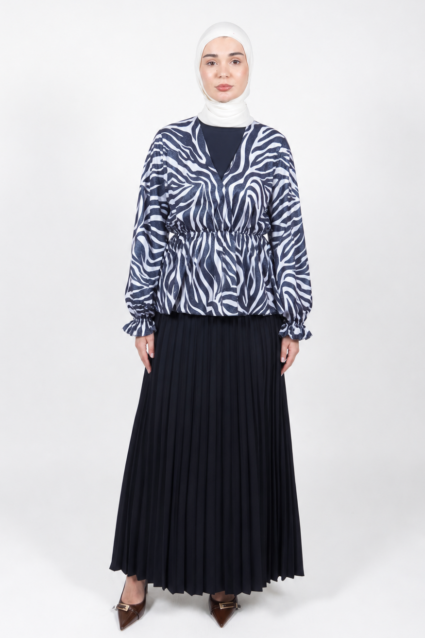 A woman modeling a long-sleeve navy and white zebra-print blouse with a V-neck and ruffled cuffs, paired with a long black pleated skirt and a white hijab, standing against a light gray studio background.