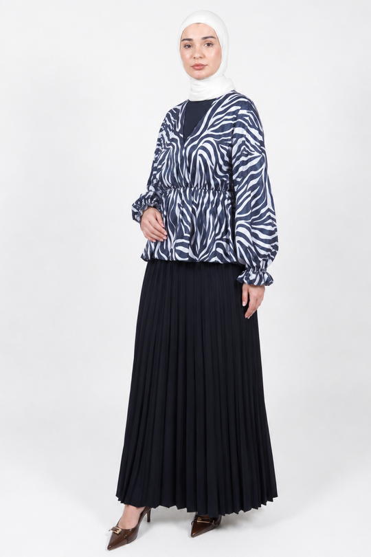 A woman modeling a long-sleeve blouse with a navy and white zebra print, paired with a long black pleated skirt and a white hijab, standing against a light gray studio background.