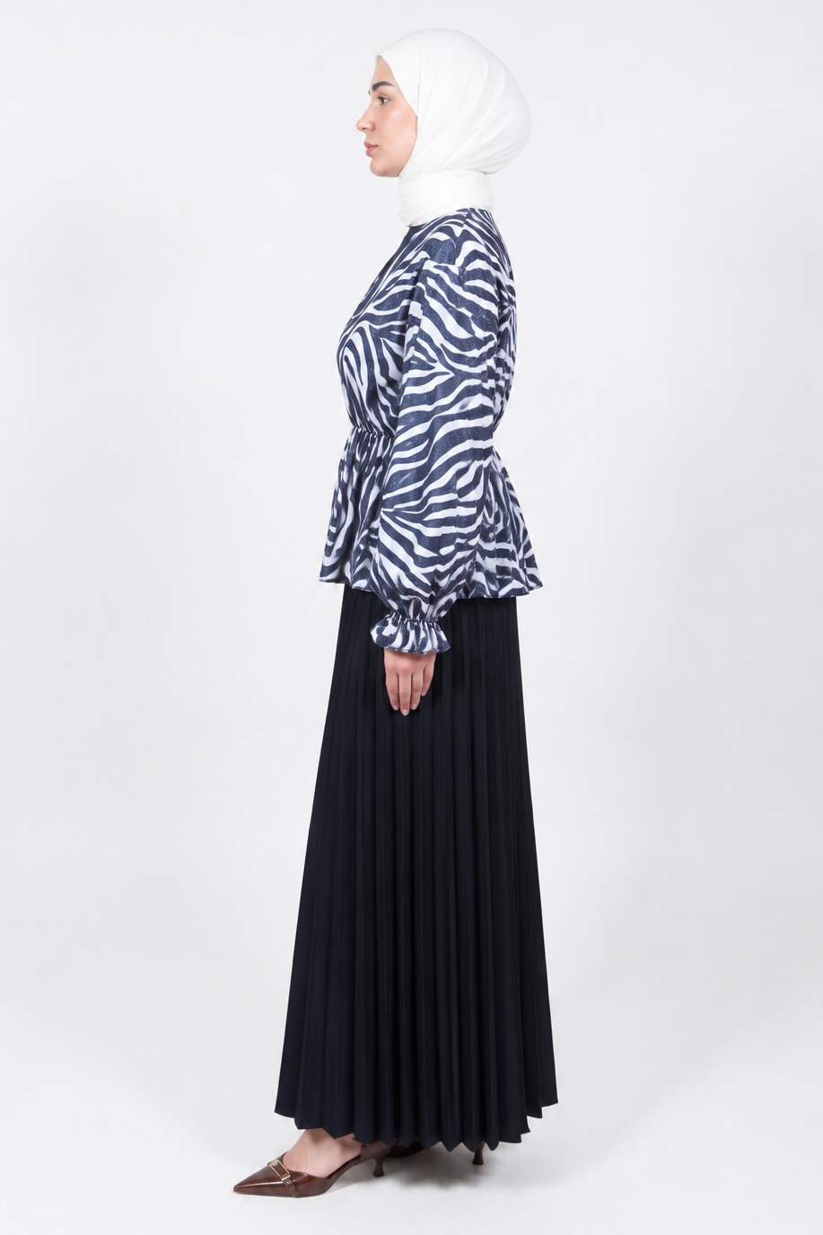A woman in profile wearing a white hijab, a navy and white zebra-print long-sleeve peplum blouse, and a long black pleated skirt, standing against a light gray studio background.