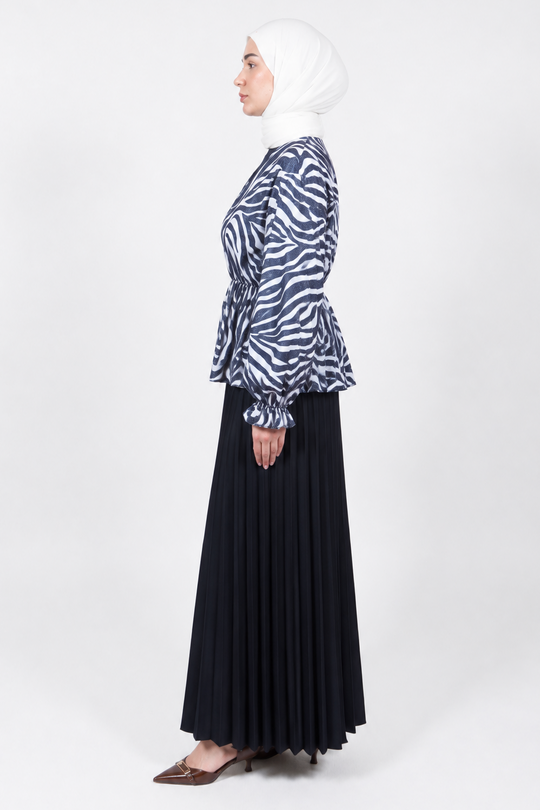 A woman in profile wearing a white hijab, a navy and white zebra-print long-sleeve peplum blouse, and a long black pleated skirt, standing against a light gray studio background.
