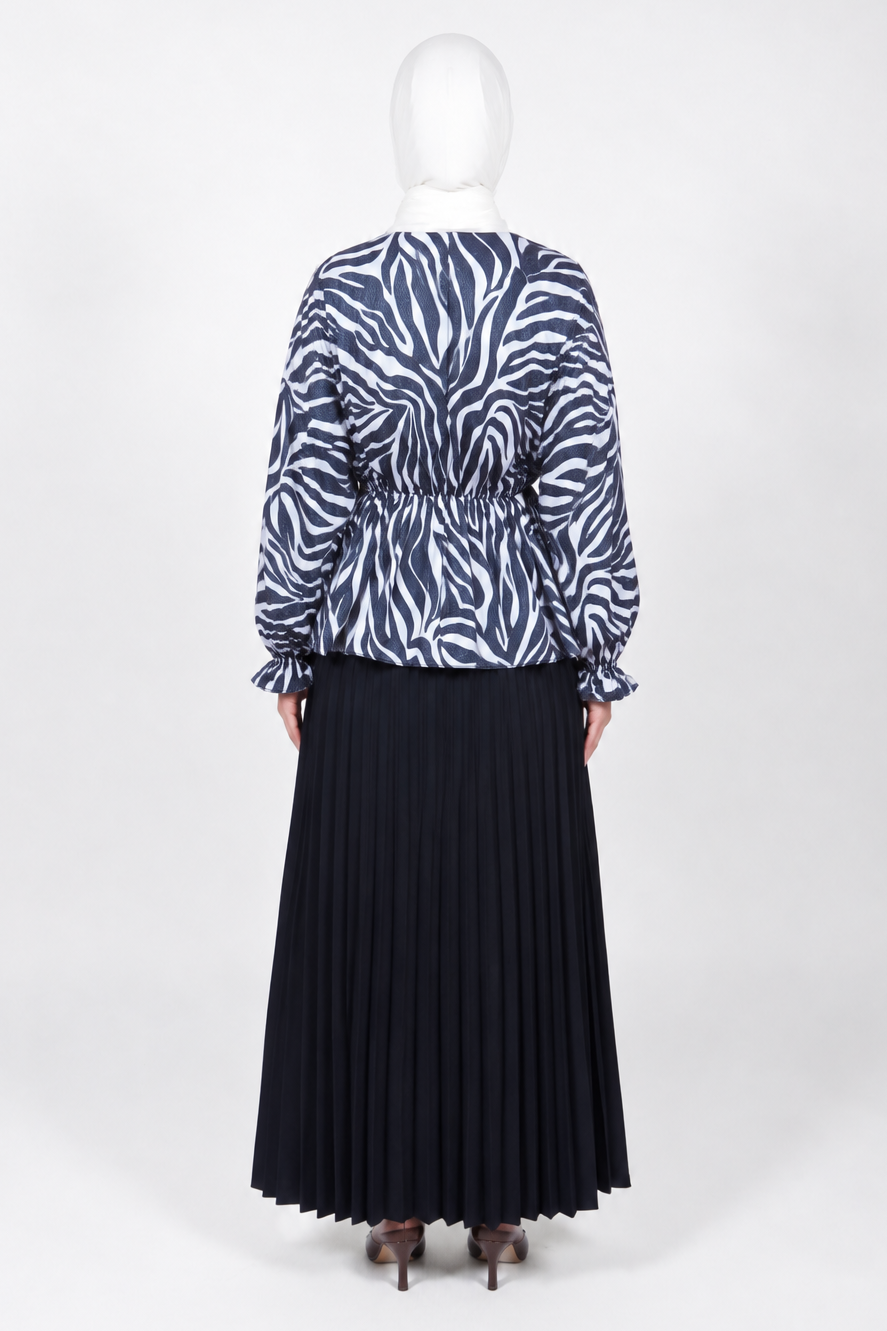 A woman stands with her back to the camera wearing a long-sleeve top with a navy and white zebra print and a long pleated navy skirt, standing against a plain white studio background.