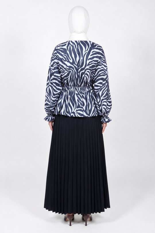 A woman stands with her back to the camera wearing a long-sleeve top with a navy and white zebra print and a long pleated navy skirt, standing against a plain white studio background.