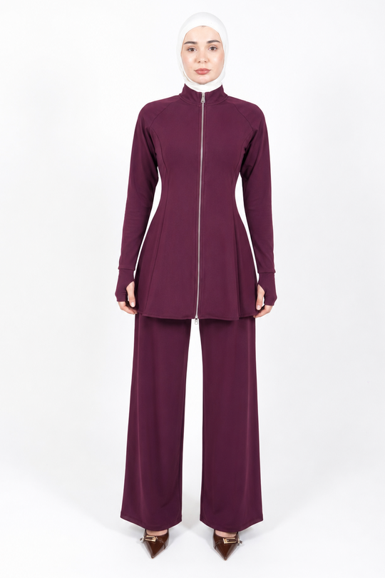 A woman modeling a matching two-piece modest outfit in deep plum purple, featuring a long-sleeve zip-front top and wide-leg pants, paired with a white hijab and brown pointed shoes, against a plain white studio background.