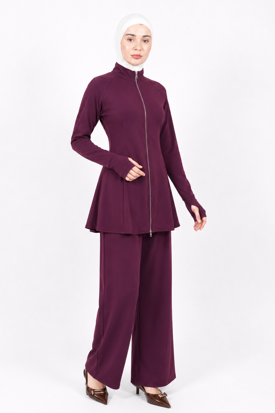 A woman modeling a matching two-piece modest outfit in deep plum purple, featuring a long-sleeve zip-up top with a high mock neck and wide-leg pants, paired with a white hijab and brown pointed-toe heels, against a plain white studio background.