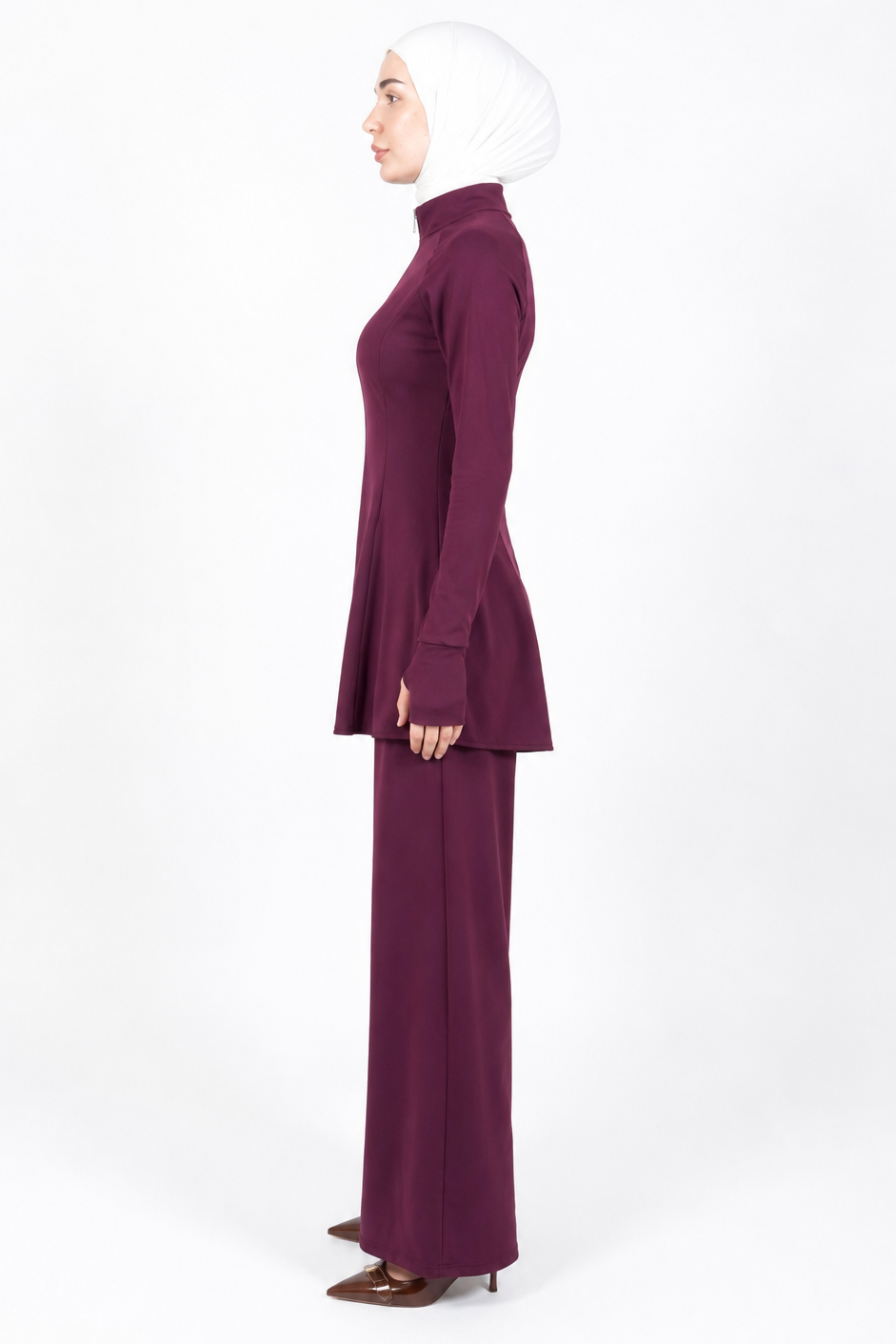 A woman in profile wearing a matching deep plum two-piece outfit with a long-sleeve mock-neck top and wide-leg pants, paired with a white hijab and brown pointed-toe heels, against a plain white studio background.