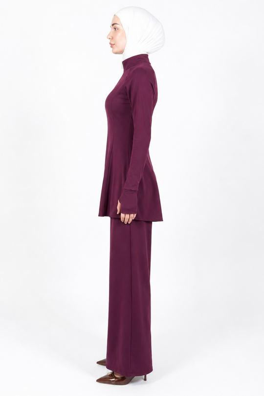 A woman in profile wearing a matching deep plum two-piece outfit with a long-sleeve mock-neck top and wide-leg pants, paired with a white hijab and brown pointed-toe heels, against a plain white studio background.