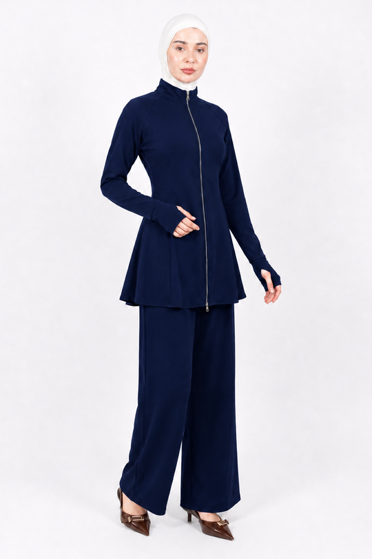 A woman modeling a navy blue two-piece modest outfit consisting of a long-sleeve zip-front top with a peplum hem and matching wide-leg pants, paired with a white hijab and brown pointed heels, against a plain white studio background.