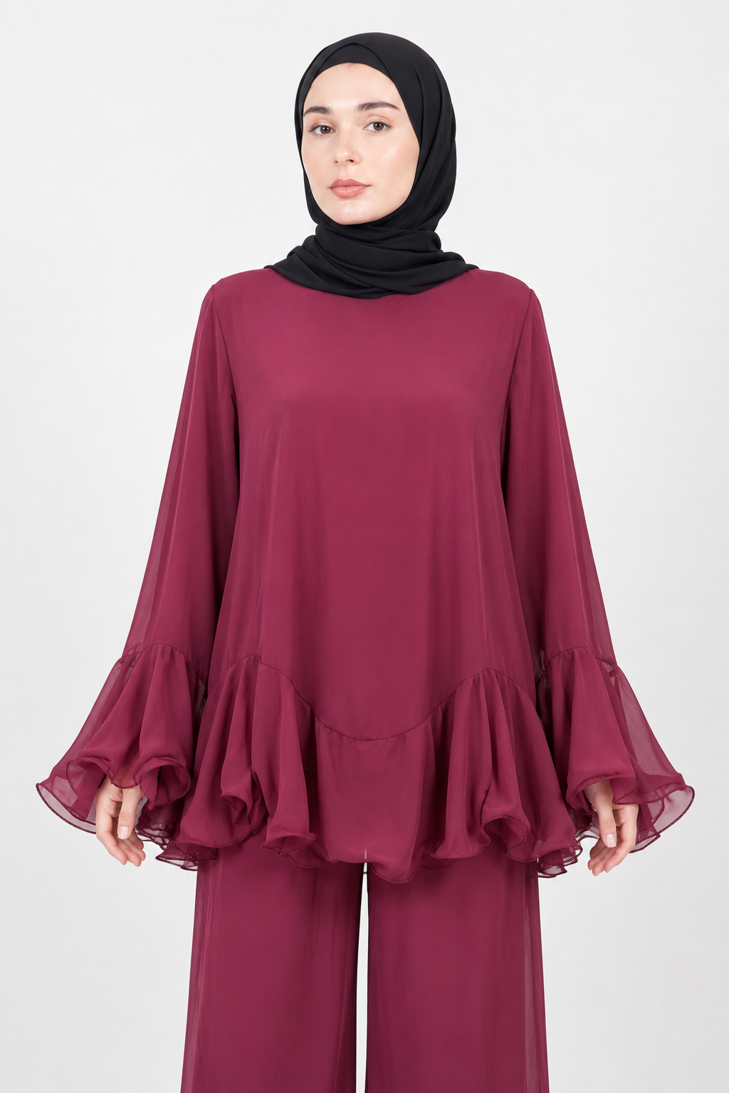A woman wearing a long-sleeved, flowy burgundy top with ruffled layers and matching wide-leg pants, paired with a black hijab, against a plain light gray studio background.