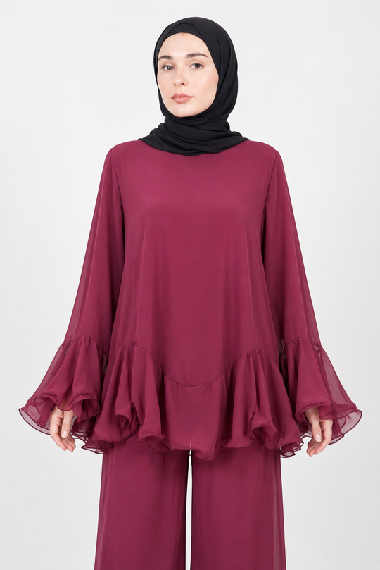 A woman wearing a long-sleeved, flowy burgundy top with ruffled layers and matching wide-leg pants, paired with a black hijab, against a plain light gray studio background.