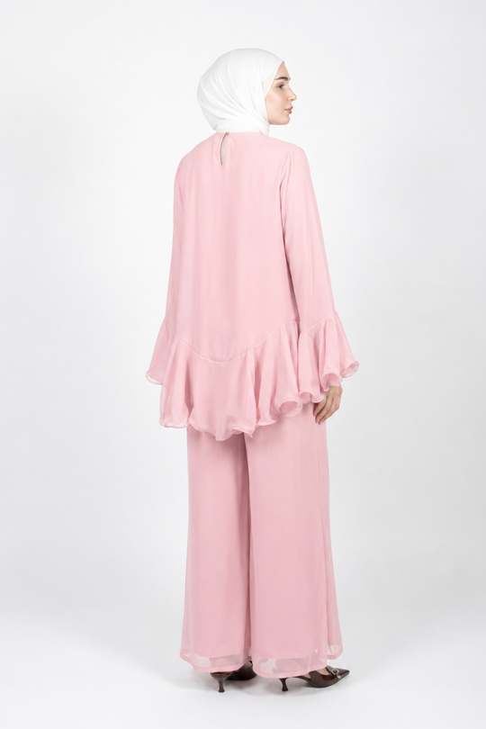A woman wearing a light pink two-piece modest outfit with a long-sleeve ruffled top and matching wide-leg pants, paired with a white hijab, shown from the back against a plain white studio background.