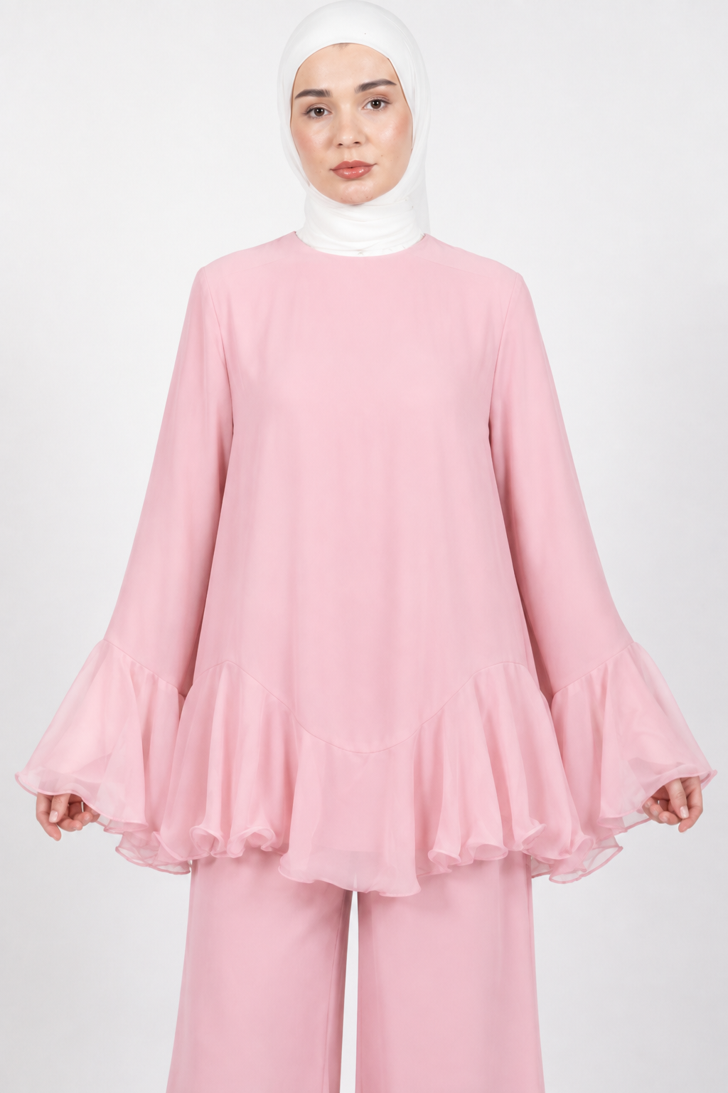 A woman wearing a light pink long-sleeve blouse with dramatic ruffled bell sleeves and matching pink wide-leg pants, paired with a white hijab, against a plain light gray studio background.