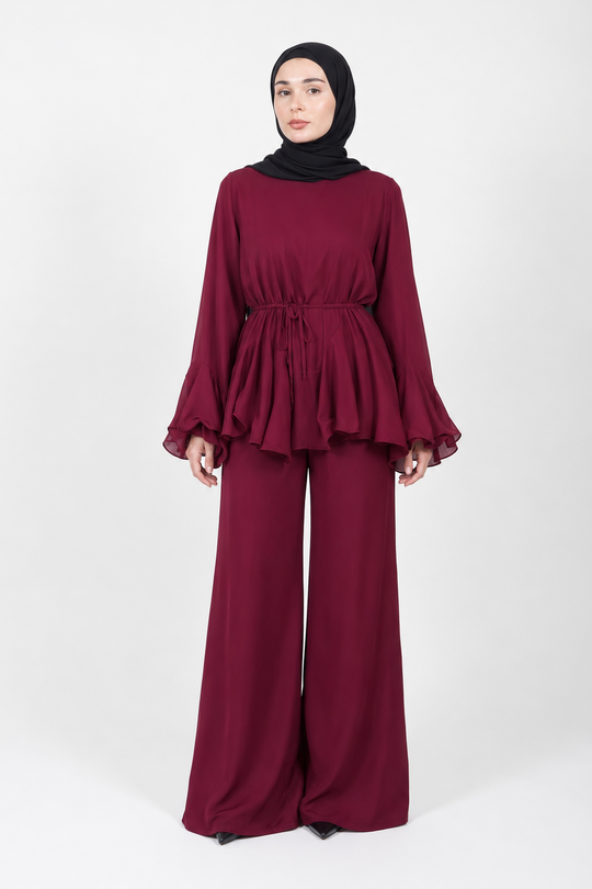 A woman wearing a matching deep burgundy two-piece outfit with a ruffled long-sleeve top and wide-leg pants, paired with a black hijab, standing against a plain light gray studio background.