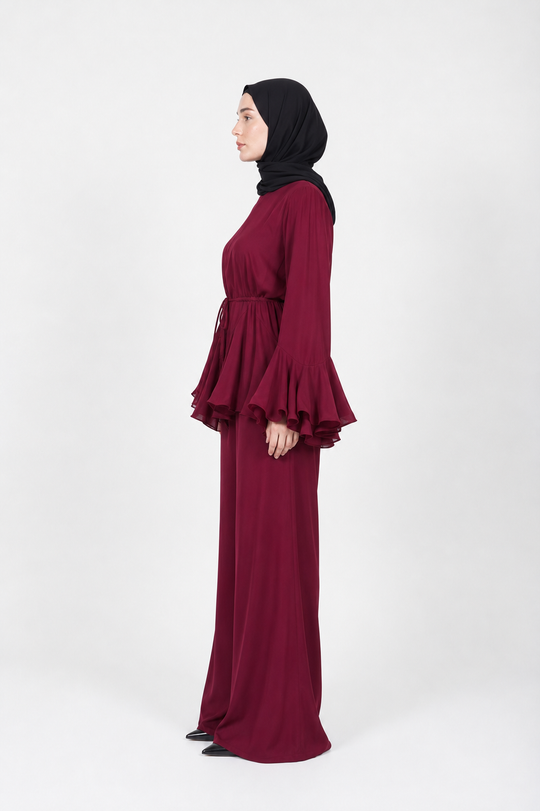 A woman in profile wearing a long burgundy maxi dress with layered ruffle sleeves and a matching black hijab, standing against a plain light gray studio background.