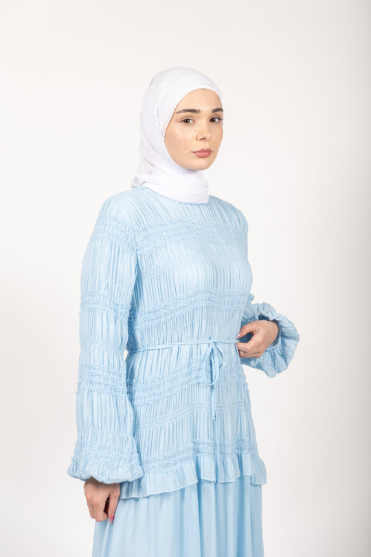Woman wearing a light blue pleated dress and white hijab on a white background
